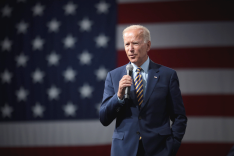 Joe Biden steht mit einem Mikrofon in der Hand vor einer großen Flagge der USA.