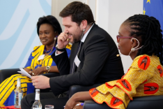 Zwei traditionell afrikanisch gekleidete Frauen und ein geschäftlich gekleideter Mann sitzen gemeinsam auf einem Podium.