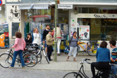 Vor einem Eckladengeschäft mit einer alten Straßenlaterne und Plasterstraße davor stehen und gehen zahlreiche Menschen verschiedener Herkunft und verschiedenen Alters, mehrere mit Fahrrädern; die gesamte Szene mutet durch Graffiti rauh und bunt an.