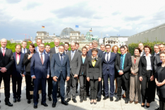 Eine Gruppe geschäftlich gekleideter Menschen steht auf einer Dachterrasse; im Hintergrund sind der Reichstag und das Brandenburger Tor in Berlin erkennbar.