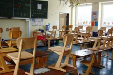 In einem Klassenzimmer stehen zahlreiche kleine Stühle und Tische aus Holz; an der Stirnseite des Raums steht eine beschriebene Tafel.