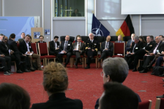 A large group of both uniformed and business-dressed people are sitting in a large circle of chairs within the Federal Academy's Historic Hall.