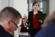 Zwei Frauen blicken zu einem General, der im Vordergrund sitzt; eine der Frauen sitzt; die andere steht, hält ein Notebook in der Hand und spricht gestikulierend.