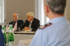 Zwei Herren im Anzug sitzen hinter Mikrofonen an einem Konferenztisch; der links im Bild blickt zu seinem Nachbarn; der rechts im Bild blickt in das Plenum.