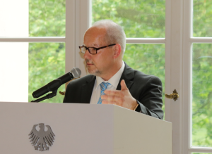 Andreas Backhoff steht vor einem Fenster mit grünem Laubwerk im Hintergrund hinter einem weißen Pult mit dem Bundesadler in Anthrazit darauf und spricht gestikulierend in ein Mikrofon. 