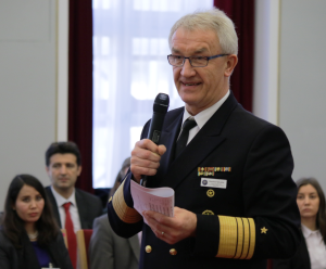 A highly decorated German Naval Officer speaks into a microphone, holding a piece of paper with notes in his hand.