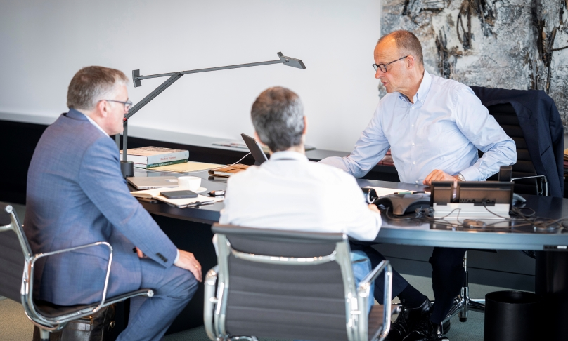 Bundeskanzler Friedrich Merz sitzt mit Regierungssprecher Stefan Kornelius und dem Abteilungsleiter 2 des Bundeskanzleramts Günter Sautter an einem Schreibtisch und spricht in ein digitales Gerät.