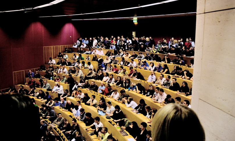 Das Bild zeigt einen sehr gut besuchten halbrunden Hörsaal, in dem alle der etagenförmig angeordneten Sitzreihen belegt sind.